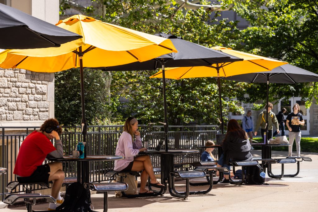 Outdoor photo of Mizzou students seated at black, metal picnic tables with alternating black and yellow umbrellas.